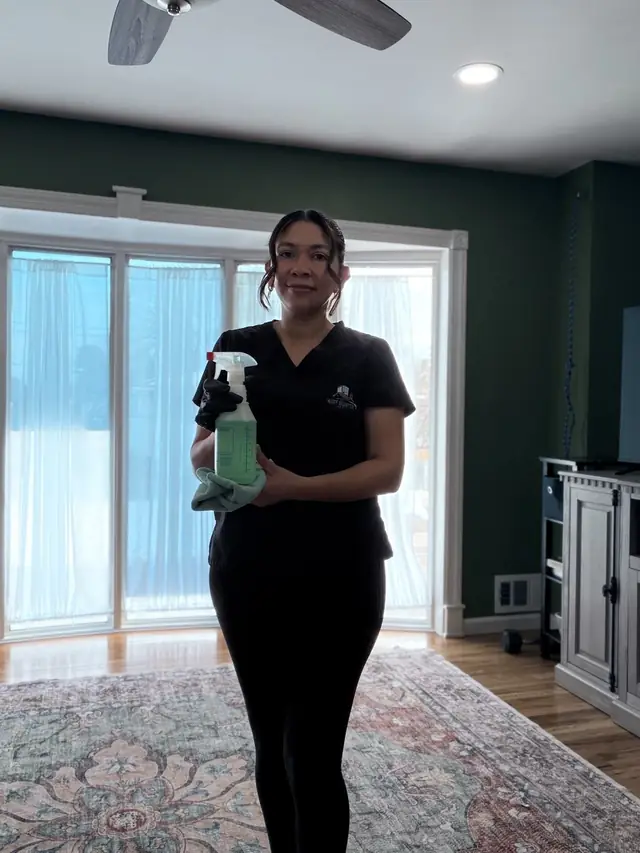 Dust Buster cleaner holding a spray bottle and cloth in a living room with a bay window